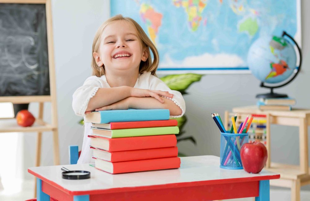 Little smiling blond girl sitting at the white desk and holding hands on the books in the spacious school classroom